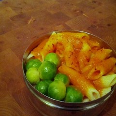 Een rond metalen bakje met daar in penne-pasta met daarboven op oranjerode saus, chilivlokken en enkele spruitjes er naast