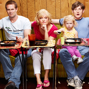 Three members of the Raising Hope family sit on sofas in front of the TV, with TV dinners on trays in front of them. The baby has comandeered the remote control. s 