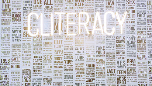 Cliteracy as a neon sign in front of a text-covered wall
