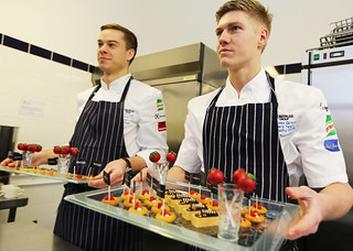 Two young men in chef aprons hold trays of canapes they've prepared.