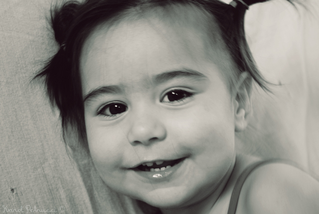 Rosto da menininha em close, ela está com a expressão alegre, sorrindo para a fotografia. A foto está em tons de preto e branco.