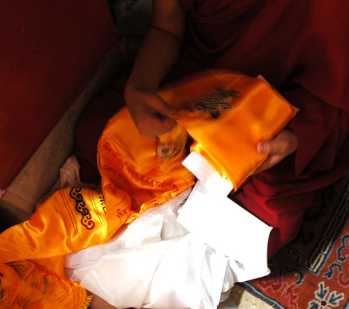 Monk prepares an offering in a envelope, white & orange decorated Tibetan khatas (ritual scarves), 8 auspicious symbols, infinity knot, seated on a Tibetan carpet, Sakya Lamdre, Tharlam Monastery of Tibetan Buddhism, Boudha, Kathmandu, Nepal by Wonderlane