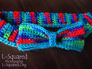 Crocheted bow tie laying on a dark wooden surface. The bow tie is made of yarn that is dyed various colors including green, teal, blue, purple and red.
