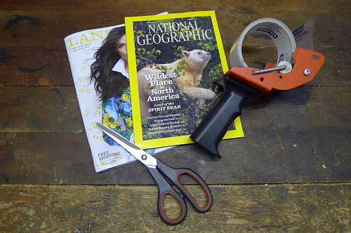 Downward view of a pair of scissors and a tape gun, sitting on top of 2 magazines, on a wood surface