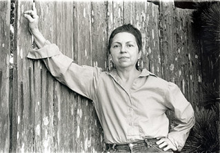 gloria anzaldua with her arm outstretched, leaning on a wooden plank fence