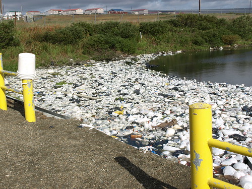 This sewage lagoon containing untreated waste,  and waste (honey) buckets convey the dire need for upgrade, along with alleviating the need to haul in drinking water.  Wind blown trash and plastic masses at the edge of the open lagoon. Photos courtesy of Larry Yerich and Tasha Deardorff, USDA 