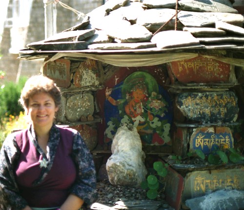 Wearing a maroon silk and poly Tibetan chuba, happy Buddhist pilgrim Linda Lane at orange Manjushri shrine, mantras carved in stone, stone roof, Dharamsala, India by Wonderlane