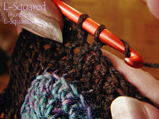 Close up of my hands as I work with brown yarn and an orange metal crochet hook; I'm in the midst of adding an edging of double  crochet stitches around a blue crocheted disk.
