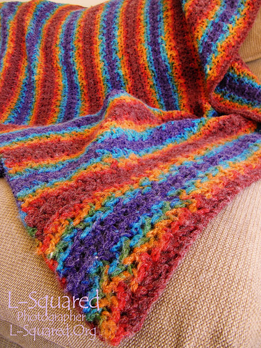 Completed blanket draped over a chair - it is made of stitches that look like tiny shells and the variegated, self-striping yarn is dark red, bright orange, school-bus yellow, bright green, turuoise and violet.
