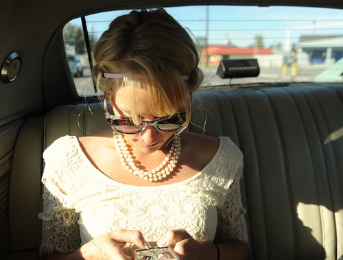 Jessie texting friends in her cream colored lace frock, sun, sunglasses and pearls, the day before the wedding, from the back seat of her fiance Chris's classic car, Fairbanks, Alaska, USA by Wonderlane