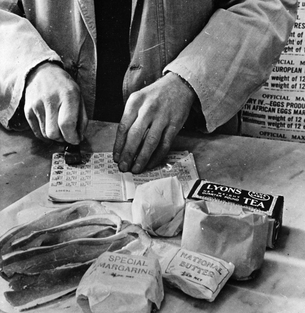 A shopkeeper cancels the coupons in a British housewife's ration book for the tea, sugar, cooking fats and bacon she is allowed for one week. Most foods in Britain were rationed and some brand names are given the designation "National".