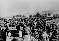 A black-and-white photo of over a hundred people in an open outdoor area. Houses are visible in the background. In the foreground there are baskets and boxes and men wearing bowler hats.
