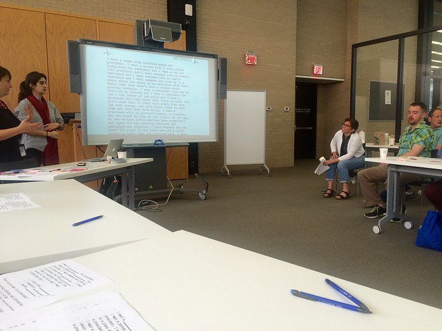 In the background, there is an electronic whiteboard with the text of a poem on it. A couple of people sit and chairs and look at the board. In the foreground, there is a white table scattered with papers and pens.