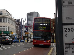 A red London bus with “West Croydon / Purley Way / Purley / 289” on the front blind, approaching along a broad road.  To the right of the photo is a foregrounded bus stop timetable for the 250 bus, and to the left are backgrounded three-story shopfronts.