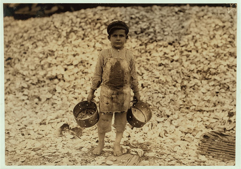 Manuel, the young shrimp-picker, five years old, and a mountain of child-labor oyster shells behind him. He worked last year. Understands not a word of English. Dunbar, Lopez, Dukate Company. Location: Biloxi, Mississippi. (LOC)