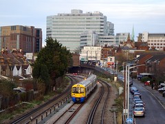 A view from above ground level showing railway tracks curving back to vanish under a high-sided bridge. Trees and houses are to the left of the line, and a road and more houses are to the right. A train with a yellow/blue back is halted on the lines. In the background, tall office blocks are visible.
