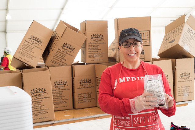 A woman holds a plastic bag of food in front of a stack of cardboard boxes.