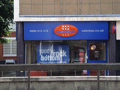 A ground-floor shopfront on the corner of a modern block.  The frontage is blue, with an oval orange sign reading “swinton” all in lower-case. Concrete barriers (part of the Croydon Underpass) can be seen in front; the photo looks to have been taken from across a busy road.