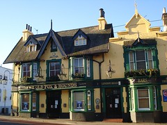 A pub with a yellow-painted frontage and dark green woodwork around the windows.  The roof has pointed gables and tall thin chimneypots.  Three sets of bay windows protrude from the front.  The name of the pub — “The Old Fox & Hounds” — is above the main entrance in gold-coloured capital letters.