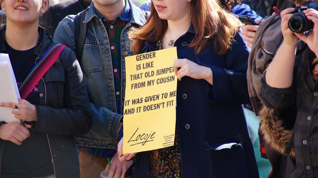 a photo of a rally where a person holds a sign saying "gender is like that old jumper from my cousin, it was given to me and it doesn't fit."
