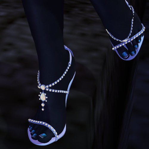 Image Description: Close up of black feet with teal toenails wearing white, jeweled sandals.