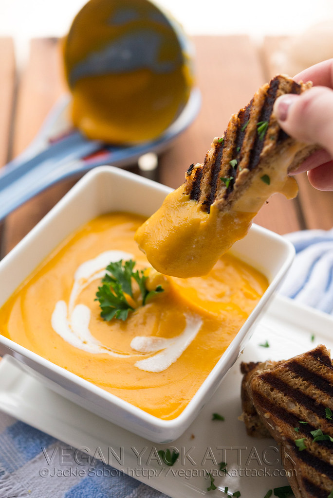 Square white bowl with bright orange butternut bisque inside, with grilled cheese stick dipped in