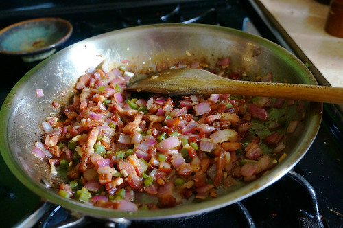 A wooden spatula lies across a skillet full of bacon and seasoning vegetables